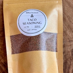 May include: A tan paper pouch containing taco seasoning with a clear window showing the brown spice blend. The circular white label reads BBQ-licious, TACO SEASONING, 2 oz/56g, barbecue seasoning, and made with love, featuring a small fire icon.