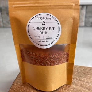 May include: A tan stand-up pouch containing BBQ-licious Cherry Pit Rub barbecue seasoning. The circular white label features a fire icon and the text 2 oz/56g. A clear window reveals the coarse, reddish-brown spice blend inside the packaging.