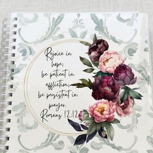 May include: A spiral-bound notebook featuring a light green damask pattern background. A circular gold frame contains the black text: Rejoice in hope; be patient in affliction; be persistent in prayer. Romans 12:12, accented by dark pink and light pink peonies.