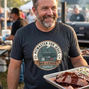 May include: Dark grey t-shirt with a circular graphic that reads "PITMASTER FOR JESUS BBQ-LICIOUS". The graphic includes a smoker illustration, cross, and fish symbol. The shirt is worn by a person holding a tray of BBQ.