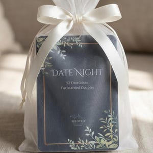 May include: A deck of cards titled DATE NIGHT 52 Date Ideas For Married Couples with a dark blue background, gold border, and green leaf illustrations, enclosed in a sheer white organza drawstring bag with a white satin ribbon bow.