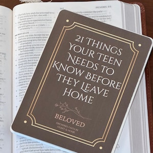 May include: A brown and gold card with the text "21 THINGS YOUR TEEN NEEDS TO KNOW BEFORE THEY LEAVE HOME" is placed on top of an open book. The card also includes the word "BELOVED" and the phrase "GROWING FAITH IN HEART & HOME."