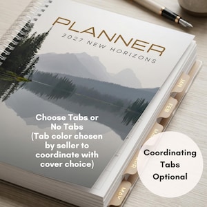 May include: A spiral-bound 2027 planner titled New Horizons featuring a landscape illustration of a lake and mountains on the cover. The planner includes monthly tabs labeled JAN, FEB, MAR, APR, and MAY along the side, with a pen resting nearby.