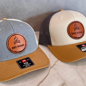 Puede incluir: Dos gorras tipo trucker con viseras color canela y parches circulares de cuero con un gráfico de fogata y el texto BBQ-licious. La gorra izquierda tiene un panel frontal gris jaspeado, la derecha tiene un panel frontal blanco hueso y malla gris.