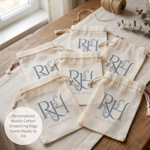 May include: A collection of off-white muslin cotton drawstring bags featuring a blue printed monogram of the letters RH intertwined with floral details and the text RAMA & HELENA. The bags are arranged in a group, each with a knotted cord closure.
