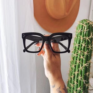 May include: Black square-framed eyeglasses with clear lenses. The glasses have a thick frame and are held up against a background with a cactus and a tan cowboy hat.