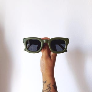 May include: Olive green sunglasses with dark lenses are held up against a white background. The sunglasses have a thick, square frame. The arm of a person with a tattoo is visible.