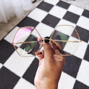 May include: Gold-rimmed, hexagonal eyeglasses with clear lenses. The glasses are held up against a black and white checkered background. The frame reads "NICKEL SILVER FRAME" and "48231CL CHINA CE".