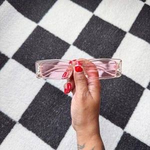 May include: Clear pink rectangular eyeglasses. The glasses have a transparent frame and lenses. The background is a black and white checkered pattern.