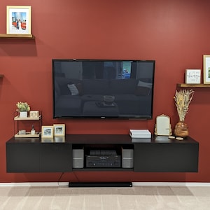 May include: A black floating entertainment center with a flat screen TV mounted above it. The wall is painted a deep red color. The entertainment center has a shelf with a stereo receiver and two speakers. There are two small shelves on the wall to the left of the TV, and a larger shelf to the right of the TV. The shelves are decorated with framed photos, plants, and decorative items.