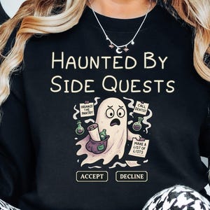 May include: Black sweatshirt with the text "HAUNTED BY SIDE QUESTS" and a cartoon ghost illustration. The ghost is holding a scroll and surrounded by speech bubbles with text. The sweatshirt has the words "ACCEPT" and "DECLINE" below the ghost.