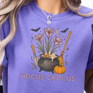 May include: Violet-colored t-shirt with a graphic featuring a cauldron, flowers, a pumpkin, and the text "Hocus Crocus." The design includes bats, a crescent moon, and gardening tools. The shirt is a comfortable, casual style.