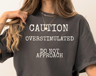 May include: Dark gray t-shirt with the text "CAUTION OVERSTIMULATED DO NOT APPROACH" in white. The shirt is worn with blue jeans and black sneakers. The overall style is casual and relaxed.