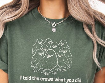 May include: Dark green t-shirt featuring a white illustration of a group of angry-looking birds. The text "I told the crows what you did" is printed below the graphic. The shirt has rolled-up sleeves.