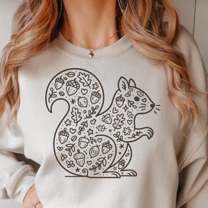May include: Beige sweatshirt featuring a black line-art squirrel design. The squirrel's body and tail are filled with acorns, leaves, and hearts. The sweatshirt has a crew neck and long sleeves, suitable for casual wear.