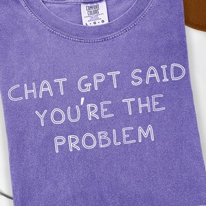 May include: A violet-colored t-shirt with the text "CHAT GPT SAID YOU'RE THE PROBLEM" in white. The shirt is folded and has a Comfort Colors tag. A brown hat is partially visible in the background.