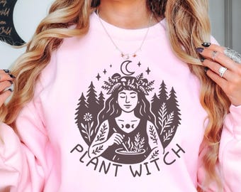 May include: A light pink sweatshirt with a graphic that says "Plant Witch". The graphic features a woman with a crescent moon and stars, surrounded by trees and plants. The sweatshirt is a crew neck style.