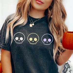 May include: Dark gray t-shirt featuring three cartoon skulls in pastel colors. The skulls are outlined in white and have a simple, friendly design. The shirt is a relaxed fit and appears to be made of soft material.