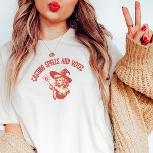May include: A white t-shirt with a red graphic of a witch casting a spell. The text on the shirt reads "Casting Spells and Votes".