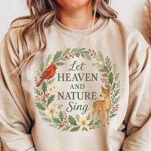 May include: Beige long-sleeve sweatshirt featuring a circular wreath design with the text "Let Heaven and Nature Sing." The wreath includes a red cardinal, a fawn, and floral elements. The sweatshirt is a neutral color.