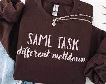 May include: A dark chocolate-colored sweatshirt with the phrase "SAME TASK different meltdown" printed in white. A silver necklace with a circular pendant is draped on the sweatshirt. The words "Dark Chocolate" are written at the bottom.