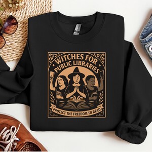 May include: Black sweatshirt with a graphic that reads "Witches for Public Libraries" in a decorative frame. The design features three figures, books, and the text "Protect the Freedom to Read". The sweatshirt is displayed with sunglasses, shoes, and denim.