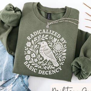 May include: Military green sweatshirt with the text "RADICALIZED BY BASIC DECENCY" in a white floral design with a bird illustration. A silver necklace is draped across the sweatshirt, which is folded on top of a pair of blue jeans.