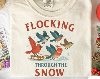 May include: Ivory-colored t-shirt with the words "FLOCKING THROUGH THE SNOW" in red. The shirt features a graphic of birds, including red cardinals and blue birds, pulling a sled. The shirt is a Comfort Colors brand.