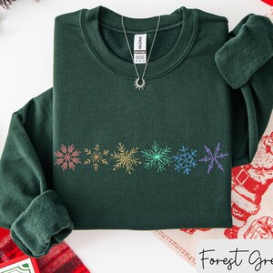 May include: A forest green sweatshirt with a crew neck and long sleeves. The sweatshirt features a row of colorful snowflake designs across the chest. A silver necklace with a sun pendant is visible. The words "Forest Green" are written in cursive.