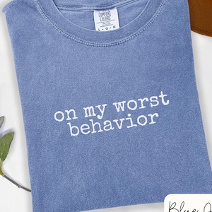 May include: A blue pigment-dyed t-shirt with a crew neckline and short sleeves. The chest features white embroidered text that reads "on my worst behavior". The interior neck label displays the brand name Comfort Colors and size L.