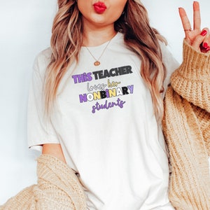 May include: A white t-shirt with the text "THIS TEACHER loves her NONBINARY students" in black, purple, and yellow lettering.