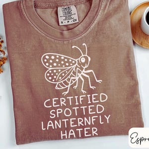 May include: Espresso-colored t-shirt with a white graphic of a spotted lanternfly and the text "CERTIFIED SPOTTED LANTERNFLY HATER." The shirt has a crew neck and a tag that reads "Comfort Colors."