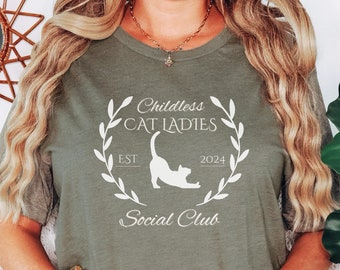 May include: Olive green t-shirt with a white graphic. The graphic features the text "Childless Cat Ladies Social Club" inside a wreath, with a stretching cat silhouette and the year "2024".