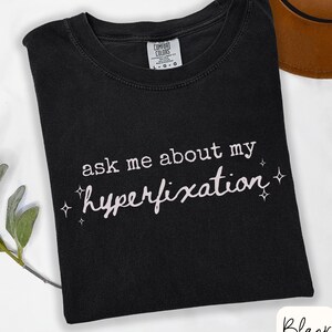 May include: Black t-shirt with the text "ask me about my hyperfixation" in white script. The shirt has a crew neck and a tag that says "Comfort Colors". A brown hat is partially visible in the background.