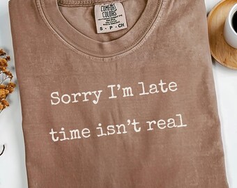 May include: A brown t-shirt with the text "Sorry I'm late time isn't real" in white. The shirt has a crew neck and a tag that says "Comfort Colors". The shirt is on a white surface with a cup of coffee and dried flowers.