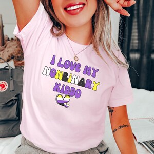 May include: Pink t-shirt with the text "I love my nonbinary kiddo" in colorful bubble letters. A purple, yellow, and white heart is below the text.