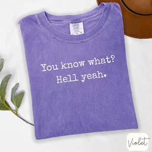 May include: A violet-colored t-shirt with the text "You know what? Hell yeah." printed in white. The shirt has a crew neck and a tag that reads "Comfort Colors". The shirt is folded and lies on a white surface.