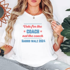May include: White t-shirt with red and blue text that reads "Vote for the *COACH* not the couch HARRIS WALZ 2024".