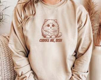 May include: A beige sweatshirt featuring a brown illustration of a fluffy cat with large eyes. The text "Cuddle Me, Bish" is printed below the cat. The sweatshirt has long sleeves and a relaxed fit.