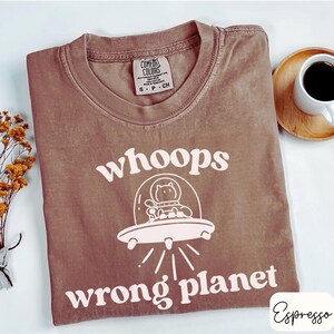 May include: Espresso-colored t-shirt with the text "whoops wrong planet" and a graphic of a cat in a spaceship. The shirt is a Comfort Colors brand, size small. A cup of coffee and a small bouquet of dried flowers are in the background.