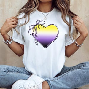 May include: White t-shirt with a purple, yellow, and white heart with a bow on top. The heart is a symbol of the non-binary gender identity.