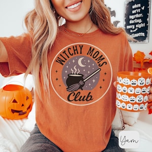 May include: Orange t-shirt with a circular graphic that reads "Witchy Moms Club." The design includes a cauldron, broom, moon, and stars. A mug with jack-o'-lantern faces is held in the hand. A carved pumpkin and a sign that says "Halloween" are in the background.