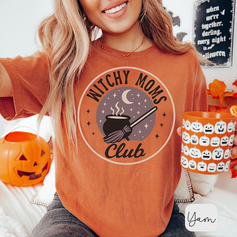 May include: Orange t-shirt with a circular graphic that reads "Witchy Moms Club." The design includes a cauldron, broom, moon, and stars. A mug with jack-o'-lantern faces is held in the hand. A carved pumpkin and a sign that says "Halloween" are in the background.
