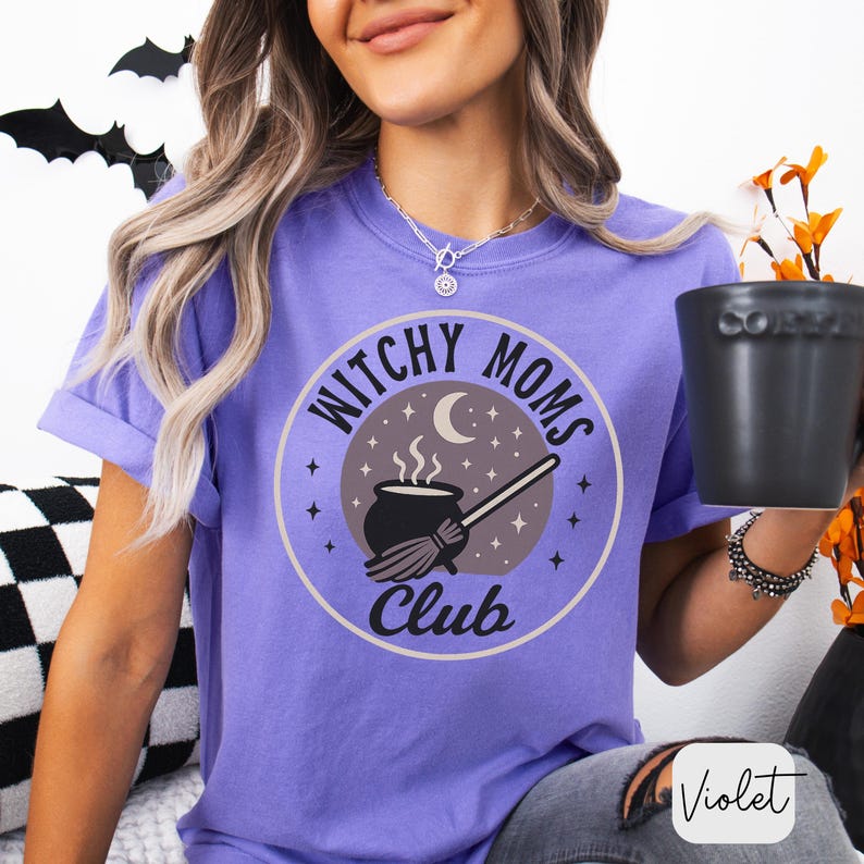 May include: A violet-colored t-shirt with a circular graphic that reads "Witchy Moms Club." The graphic features a cauldron, broom, moon, and stars. The shirt is a short-sleeved, crew neck style.