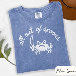 May include: Blue jean Comfort Colors t-shirt with the text "all out of spoons" and a crab graphic holding a knife. The crab has an angry expression. The shirt is folded on a white surface.