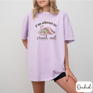 Pu&ograve; includere: T-shirt lilla chiaro con la scritta "I'm about to crash out" e un ranocchio dei cartoni animati che dorme su un cuscino. Il ranocchio &egrave; verde, e il cuscino e la coperta sono rosa. La maglietta &egrave; ampia.