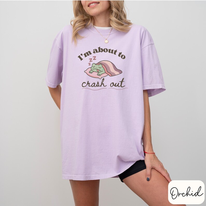 Pu&ograve; includere: T-shirt lilla chiaro con la scritta "I'm about to crash out" e un ranocchio dei cartoni animati che dorme su un cuscino. Il ranocchio &egrave; verde, e il cuscino e la coperta sono rosa. La maglietta &egrave; ampia.