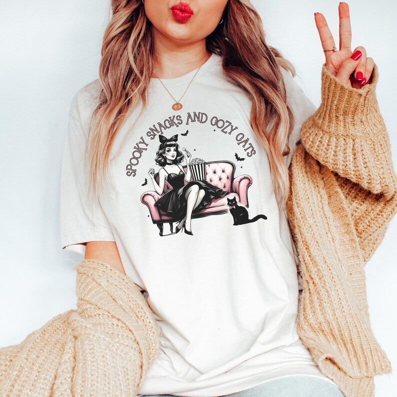 May include: White t-shirt with a graphic of a woman in a black dress sitting on a pink couch with a black cat. The text "Spooky Snacks and Cozy Cats" is written in a circle around the graphic.