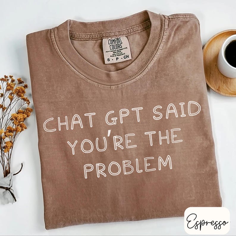 May include: A brown Comfort Colors t-shirt with the text "CHAT GPT SAID YOU'RE THE PROBLEM" in white. The shirt is laid flat on a white surface, with a small sprig of dried flowers and a cup of coffee in the background.