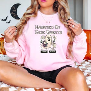 May include: A light pink sweatshirt with the text "Haunted By Side Quests" above a cartoon ghost character. The ghost holds a bag and papers with tasks. The word "Accept" and "Decline" are below the ghost.